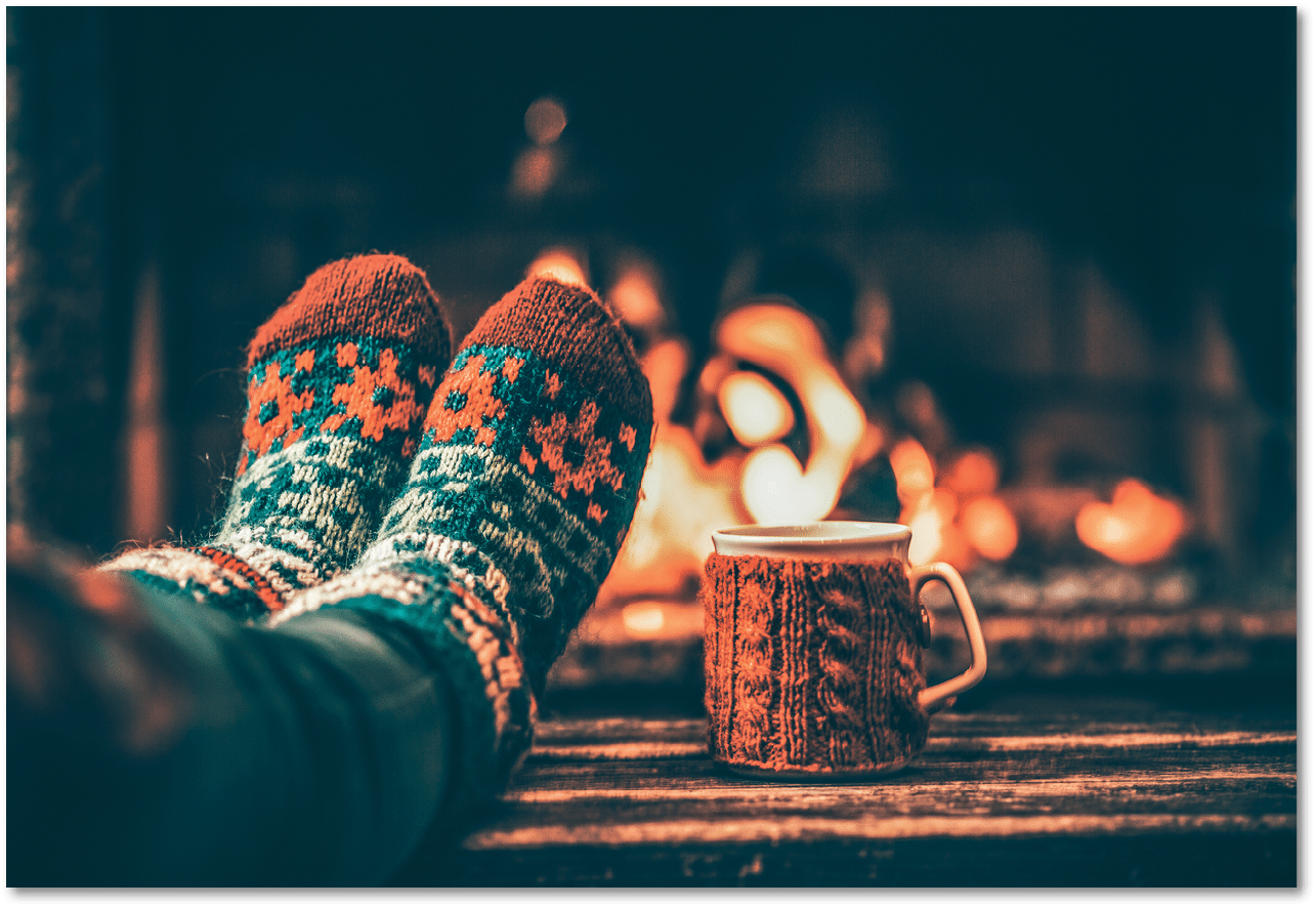 Feet in woollen socks by the Christmas fireplace. Woman relaxes by warm fire with a cup of hot drink and warming up her feet in woollen socks. Close up on feet. Winter and Christmas holidays concept.